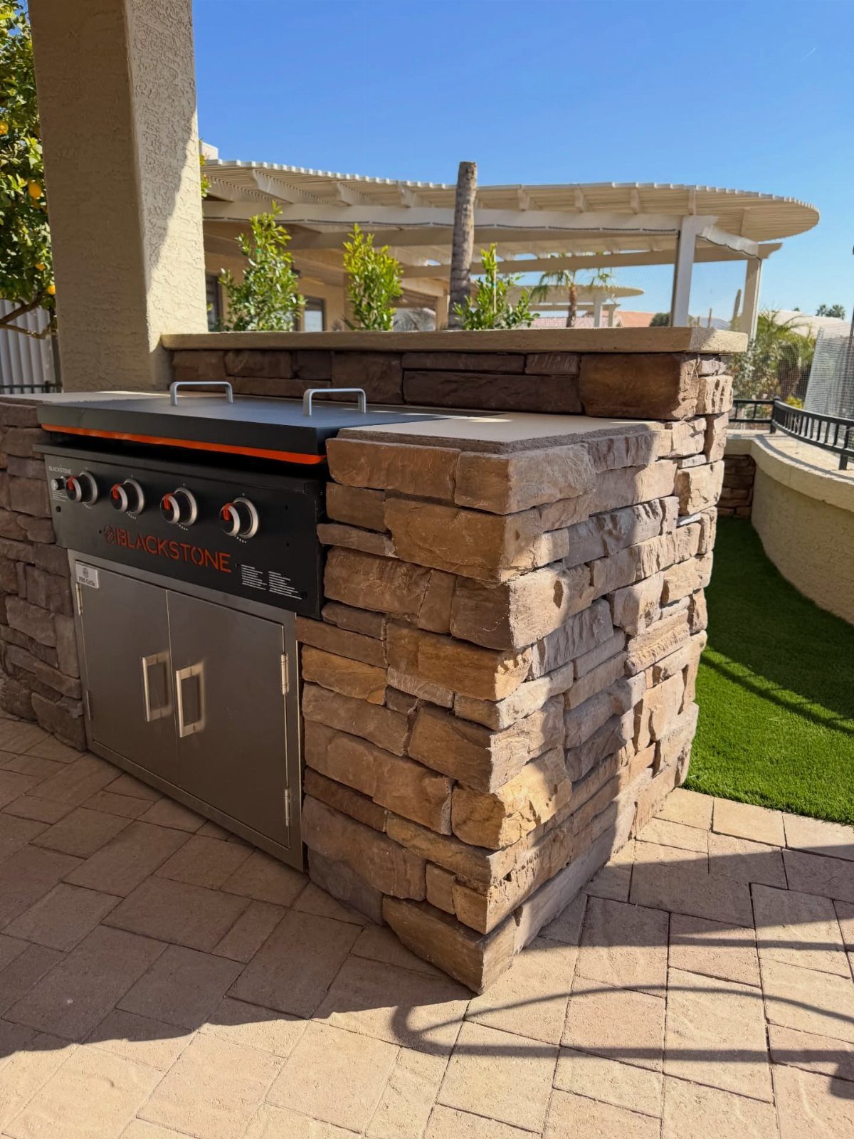 A stone outdoor kitchen island featuring a built-in stainless steel grill on a paved patio.