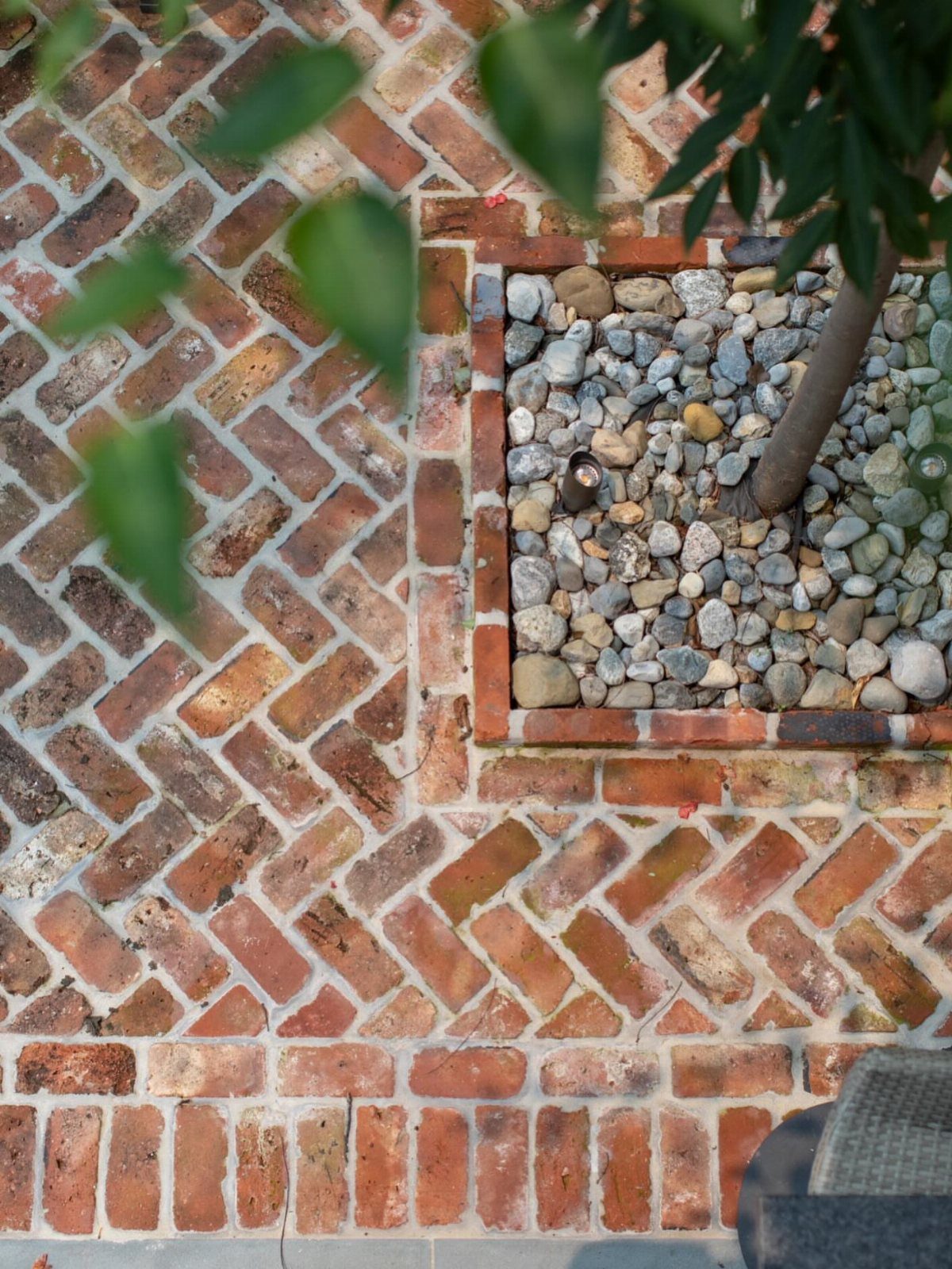 Red brick patio laid in a herringbone pattern next to a pebble-filled tree planter.