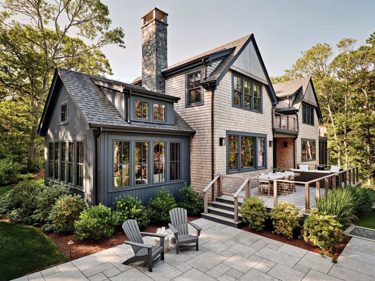 A Cape Cod style house with grey shingle siding, dark trim, and a stone chimney surrounded by lush greenery.