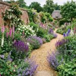 A gravel path winds through a vibrant cottage garden filled with purple, pink, and white flowers.