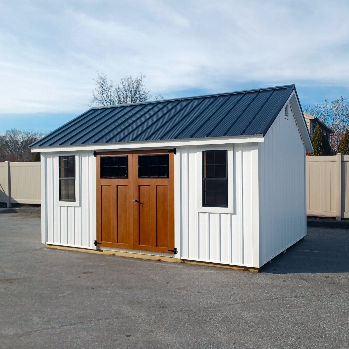 White wooden shed with a dark metal roof and brown double doors on asphalt.