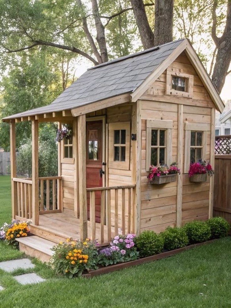 A small wooden shed house with a porch and flower boxes sitting on a green lawn.